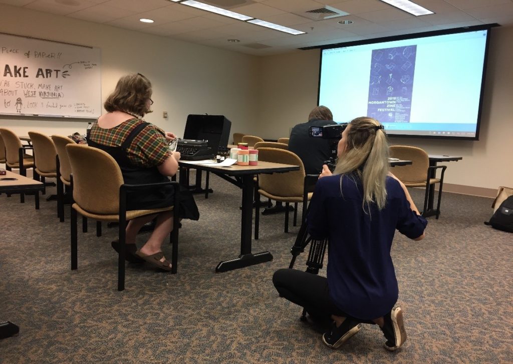 Woman working on craft project while another woman takes video.