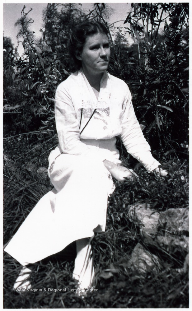 Margaret Prescott Montague in white dress, seated amid foliage in the sunshine