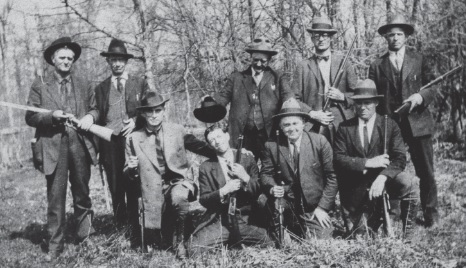 Group photo of men in suits, outdoors, most holding rifles or shotguns.