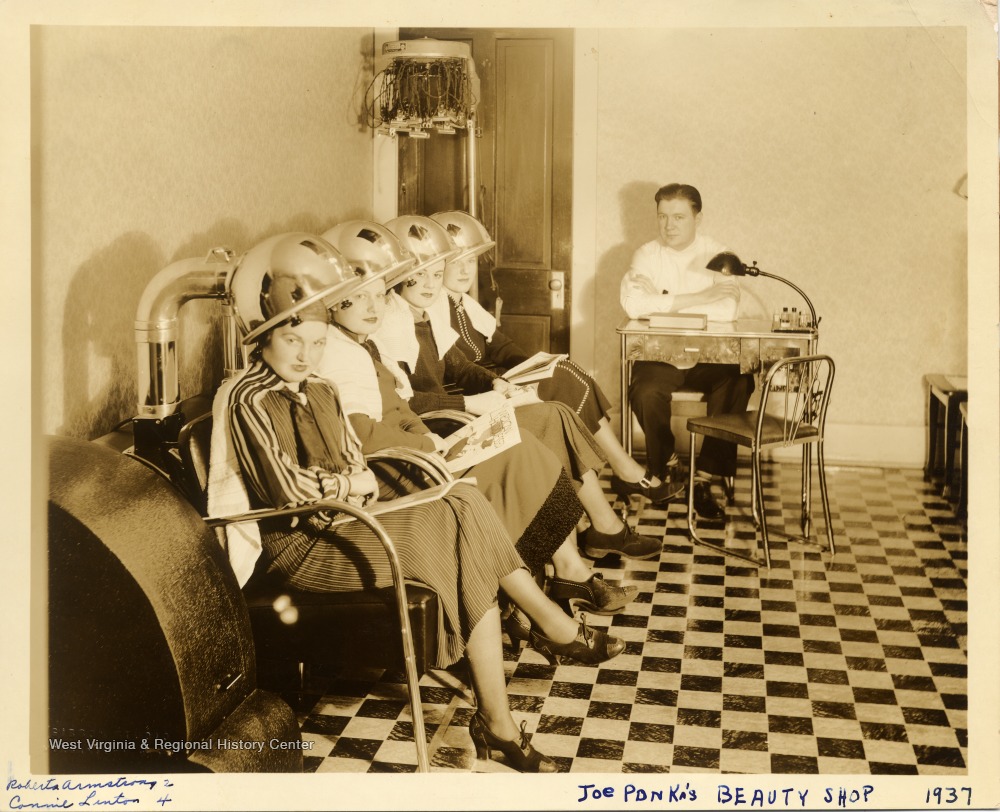 Four women under hairdryers in a salon as male staffer looks on