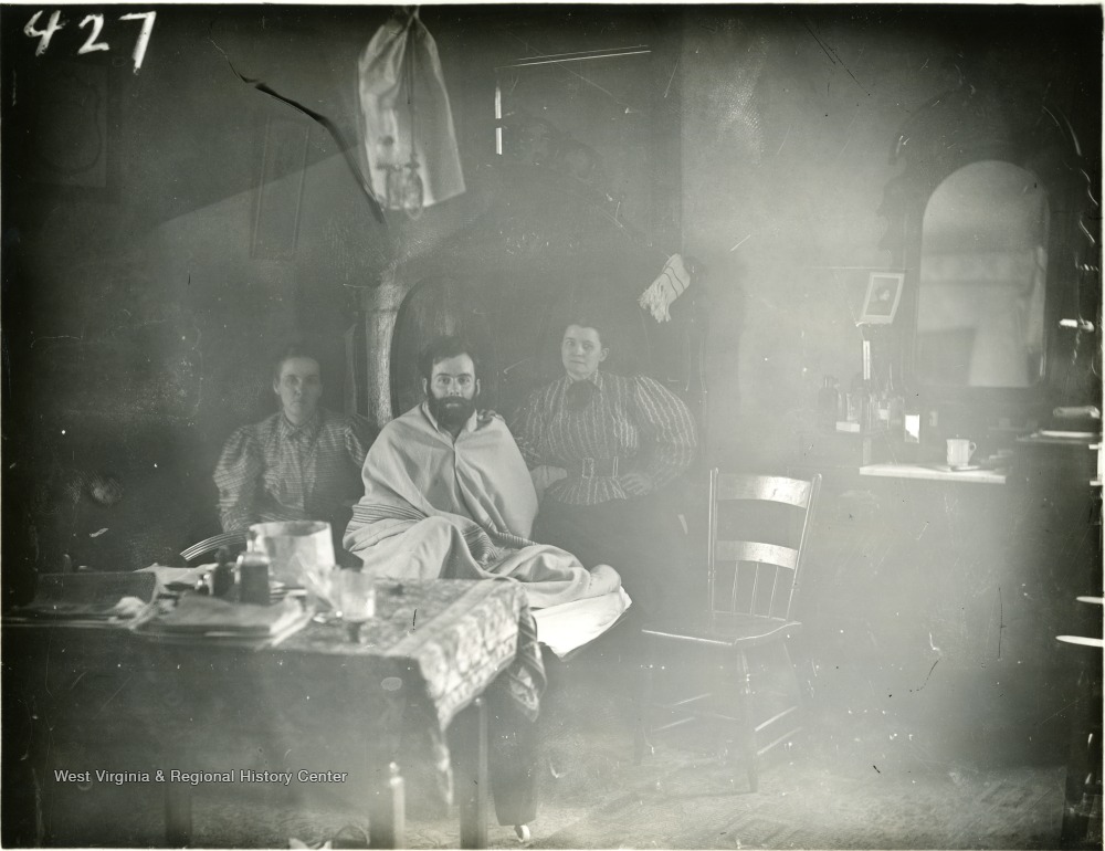 Seated man wrapped in sheet, awaiting haircut, between two women