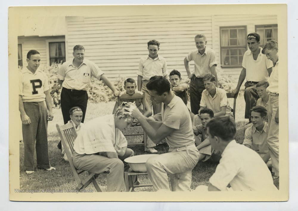 Man demonstrates washing a boy's hair over a bowl as group of boys watches outdoors