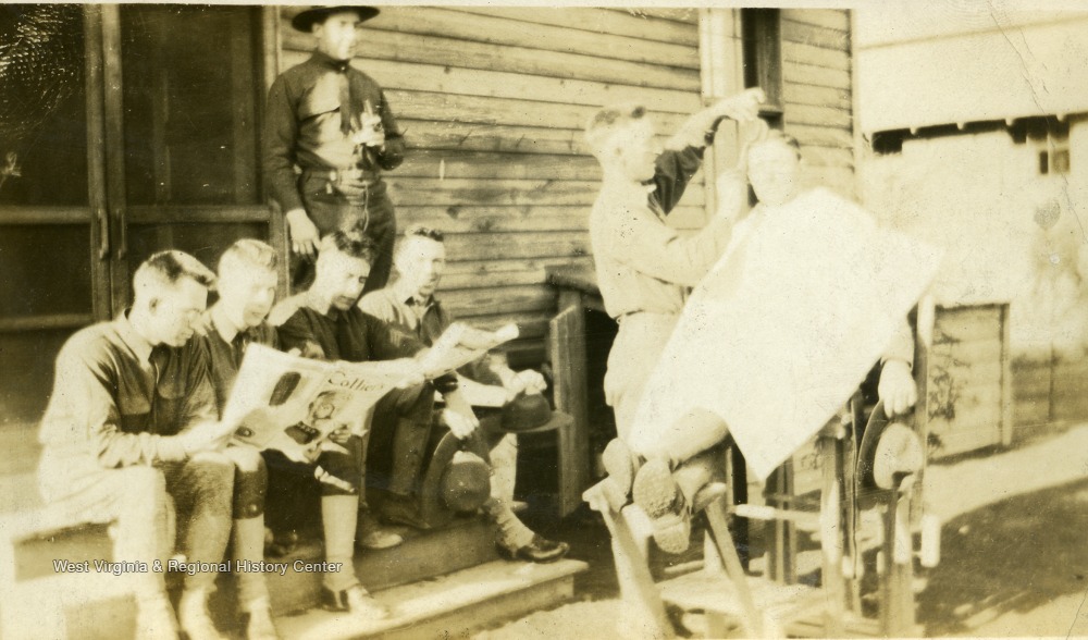 Soldiers reading newspaper outside while waiting as outdoor barber combs another soldier's hair