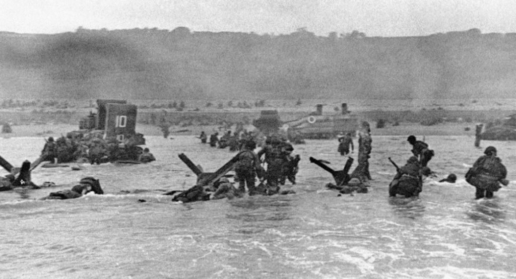 Photograph of soldiers in knee-deep water with heavy equipment, moving their way toward a beach