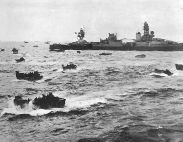 Photo of dozens of smaller boats full of people, with much larger naval vessel in the background