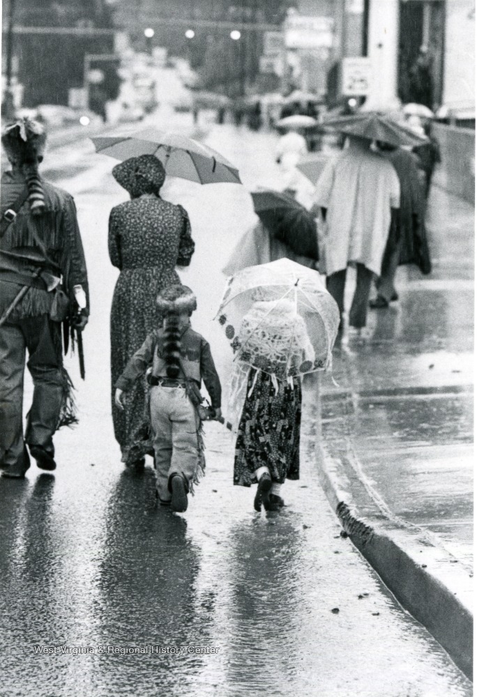 Family walking down a street in the rain, with the man and boy in Mountaineer costumes and the women in pioneer style dresses.