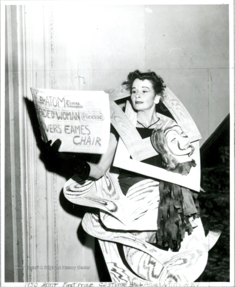 Woman in costume made of frames and a paper mache head, holding newspaper, partly obscured, that reads, "The Atom Times, __eaded woman (Picasso) ___overs Eames Chair"