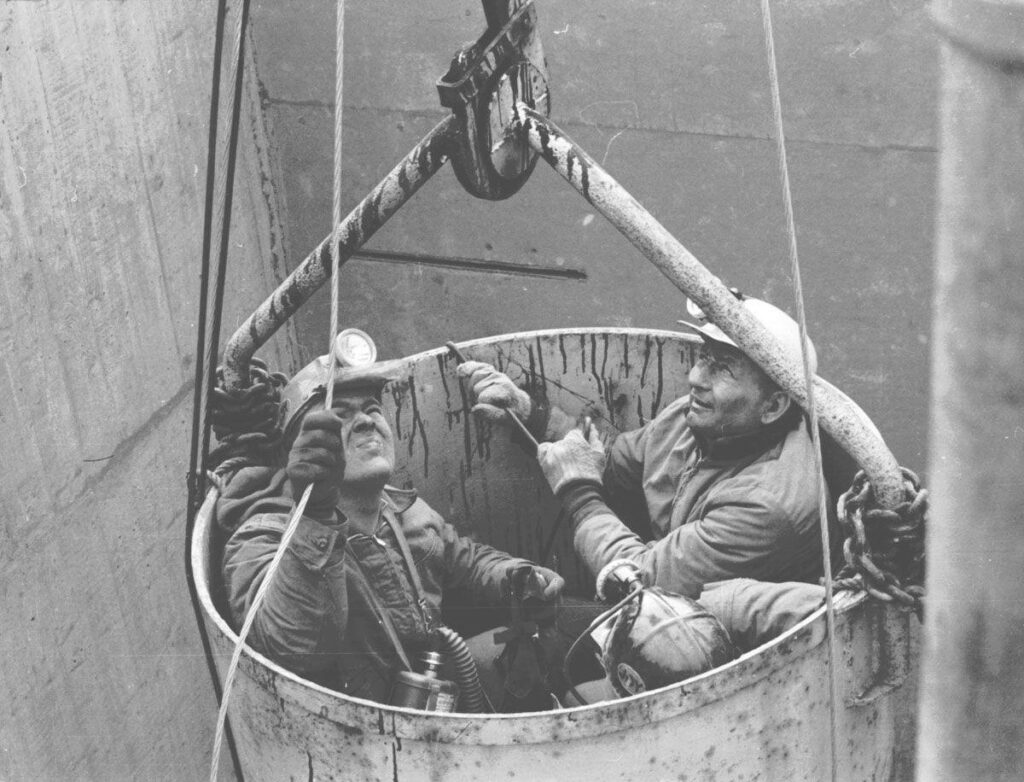 Three men in coal mining helmets, looking upwards from a bucket that is carrying them up a shaft