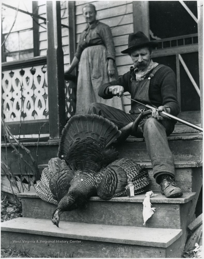 Woman standing on porch next to seated man who is cleaning his gun. A dead turkey is displayed at his feet.