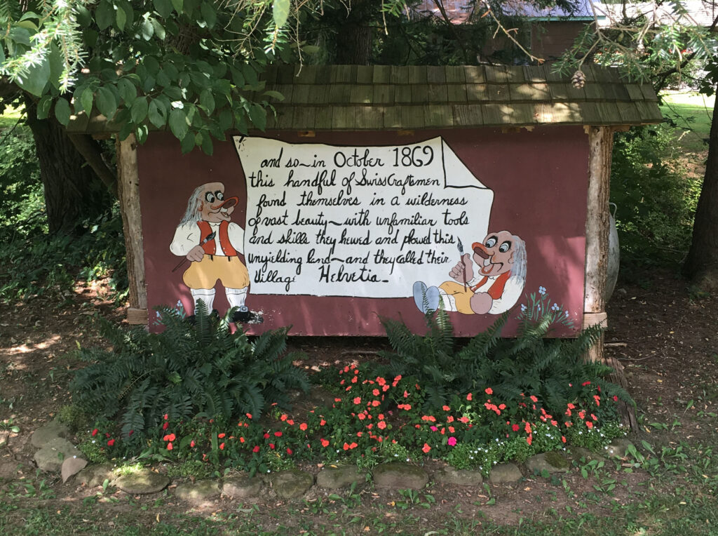 Photograph of an outdoor sign at Helvetia that reads, “and so in October 1869 this handful of Swiss Craftsmen found themselves in a wilderness of vast beauty – with unfamiliar tools and skills they hewed and plowed this unyielding land – and they called their village Helvetia.” 