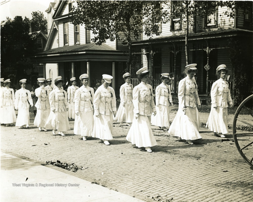 Women in white hats, jackets, and skirts march down a cobblestone street.