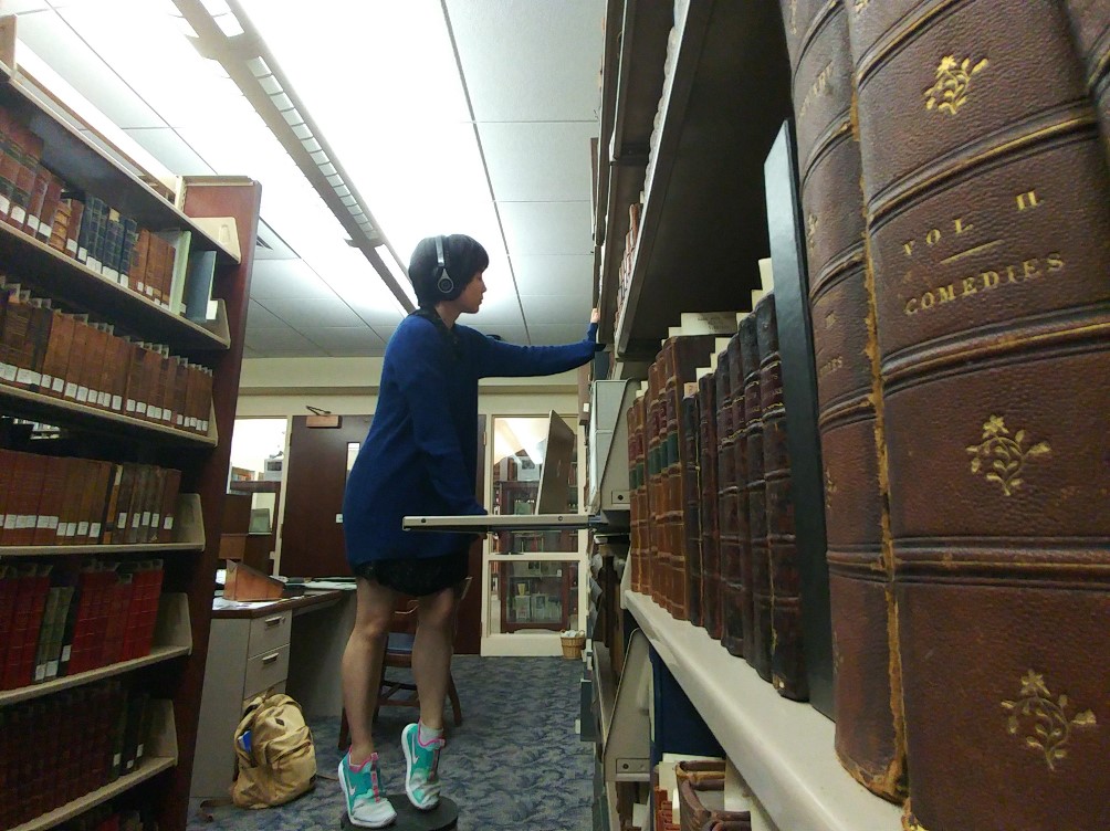 Woman between rows of bookshelves, standing on stool