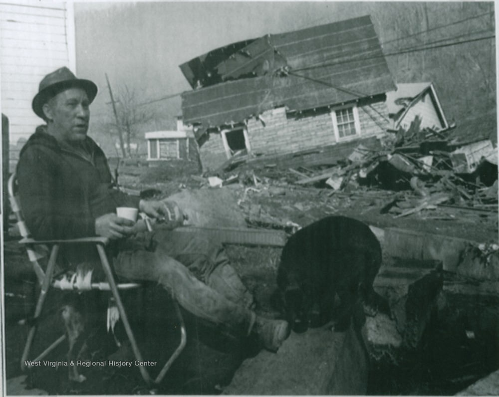 Man sitting in front of a house that has been swept off its foundation by a flood