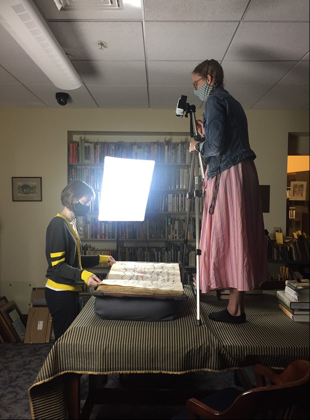 Woman operating camera on tripod to photograph a book as another woman turns pages