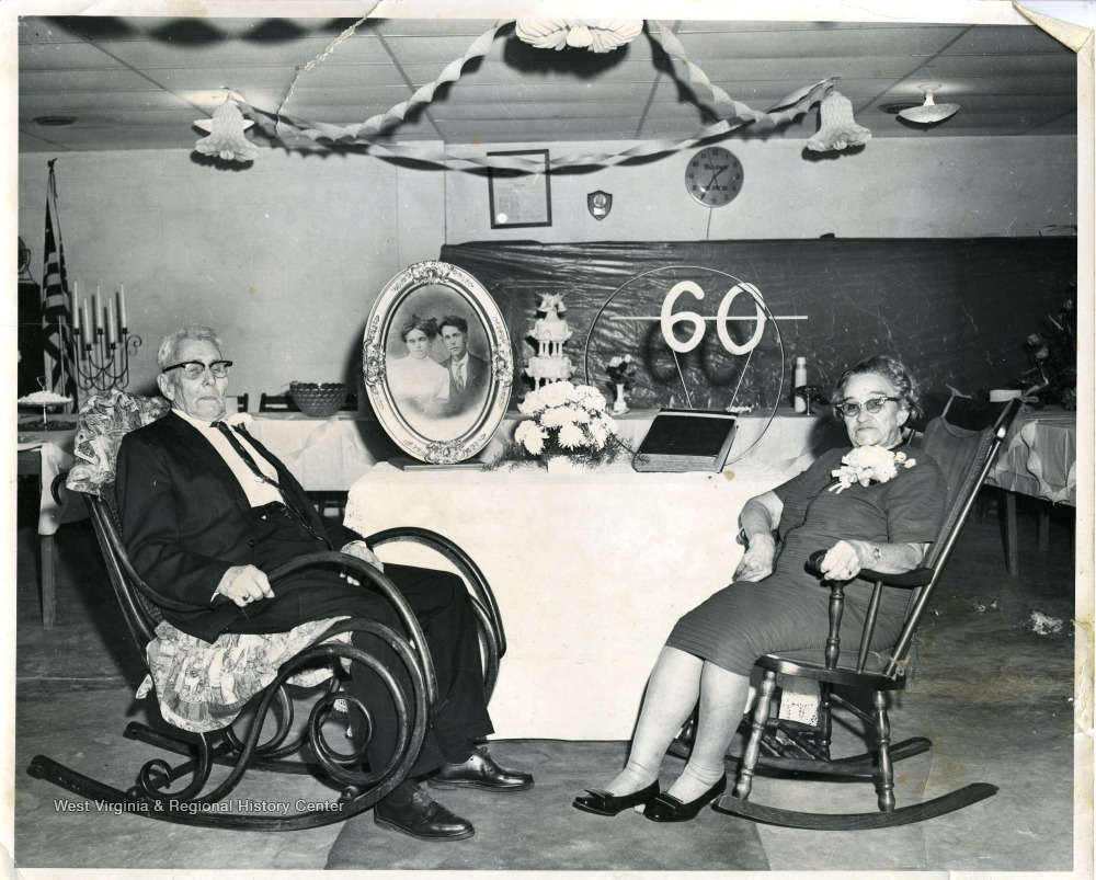 An elderly man and woman in rocking chairs, in front of an old photo and a cake, with a "60" decoration