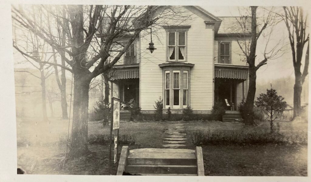 A house in black and white, surrounded by 3 trees without leaves. A short stone pathway leads to a bay window.