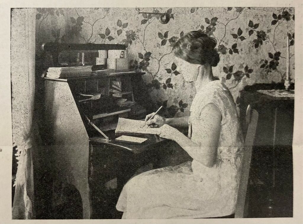 A woman in a slip dress sits at a wooden desk, holding a pencil and working on her accounts. 