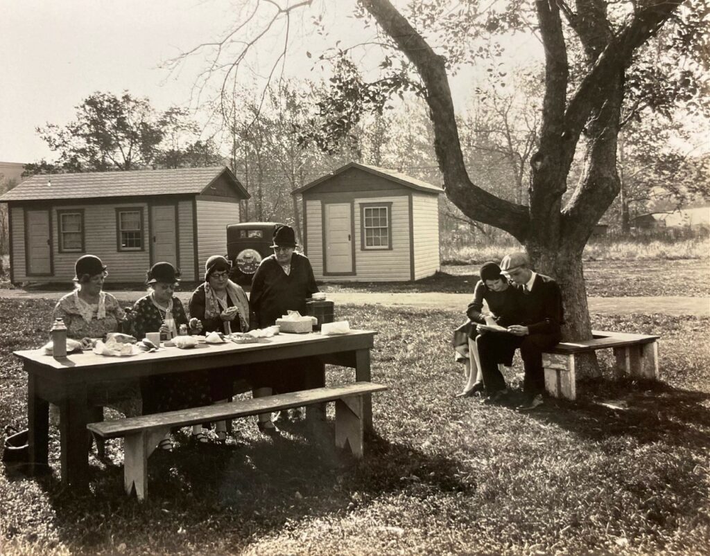 Four people sit at a picnic table outside eating breakfast. A couple sits on a bench by a tree looking at some papers.