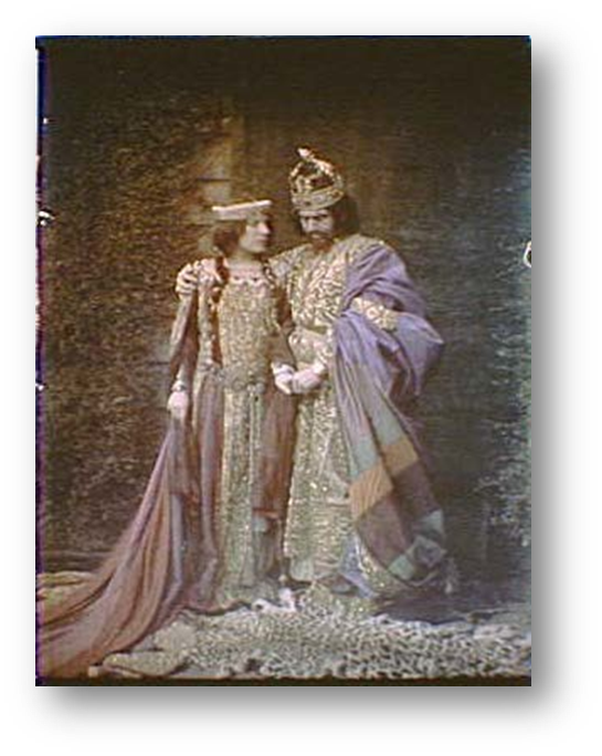A woman and a man, dressed in ornately decorated robles and dresses, each wearing golden crowns and covered in golden detailing. They hold hands, the man looking downward. 