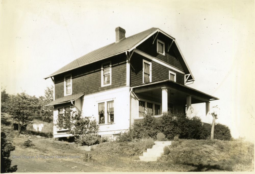 Sepia toned photo of a house with a covered porch and a barn style roof sitting a top a hill.