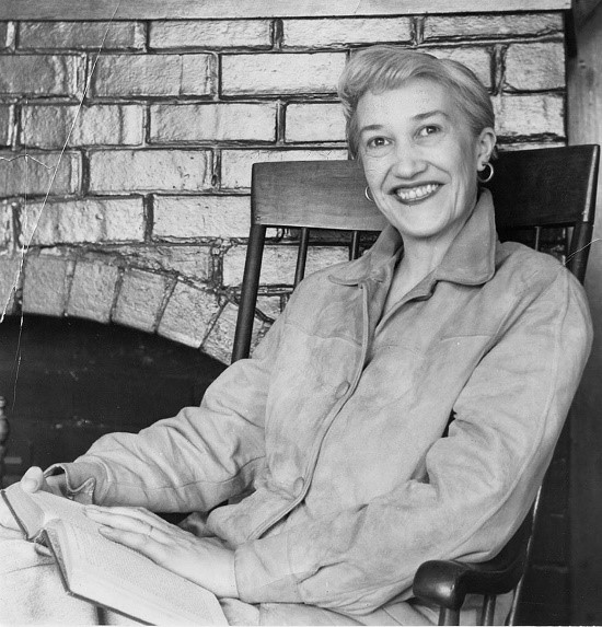 A middle aged woman with hoop earrings, styled grey hair, and a suede jacket sits in a rocking chair with a book on her lap, smiling at the camera.