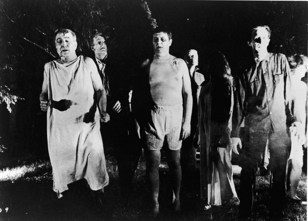 Extras from Night of the Living Dead in zombie attire walk outside.  