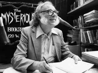 Isaac Asimov sitting at a table at The Mysterious Bookshop. He is wearing thick-framed glasses, has white sidburns, and is smiling up at someone while holding a pen.