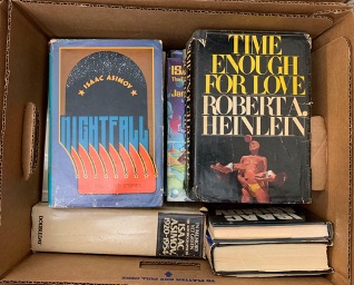 A cardboard box of rare books, the two facing upward toward the camera are "Nightfall," by Isaac Asimov and "Time Enough for Love," by Robert A. Heinlein. 